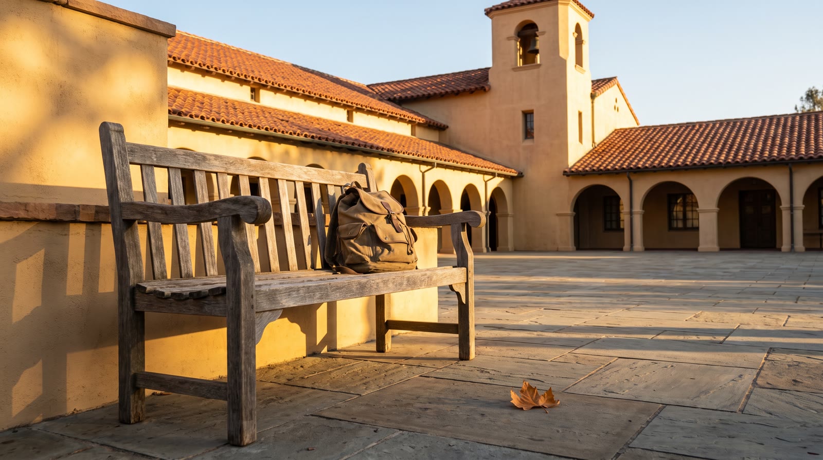 An empty wooden bench outside a Spanish-tile campus building in late afternoon light, backpack resti